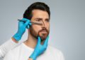 Doctor in protective gloves drawing marks on man's nose for cosmetic surgery operation, grey