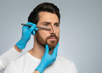 Doctor in protective gloves drawing marks on man's nose for cosmetic surgery operation, grey