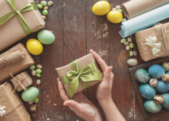 A young woman holds a gift in her hands. Colored eggs and holiday symbols on a dark wooden table. Zero waste concept.